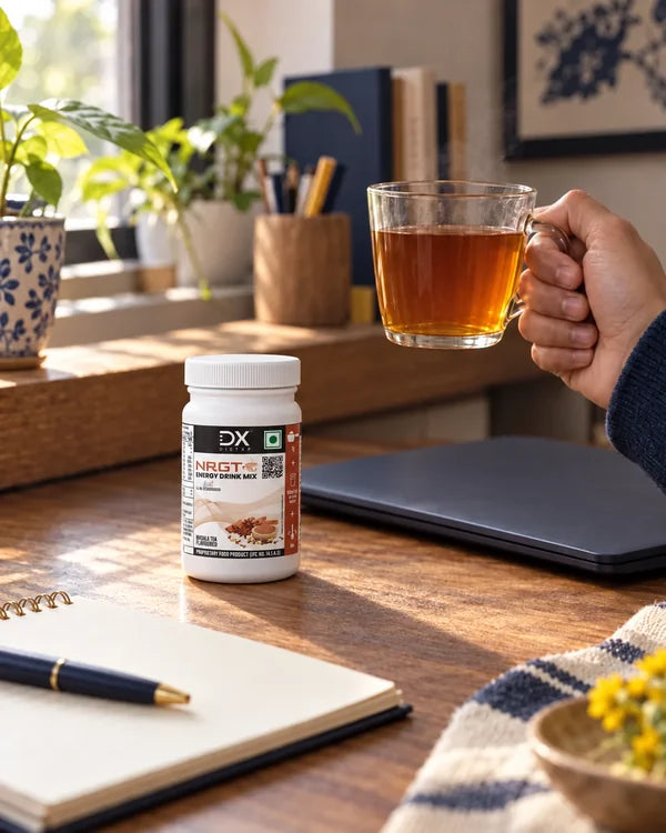 A hand holding a cup of tea beside the Masala Tea jar and a laptop
